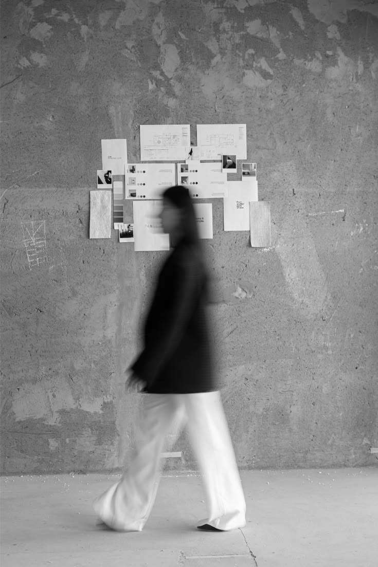 Figura difuminada caminando frente a un moodboard de diseños y muestras, pared de estudio de hormigón